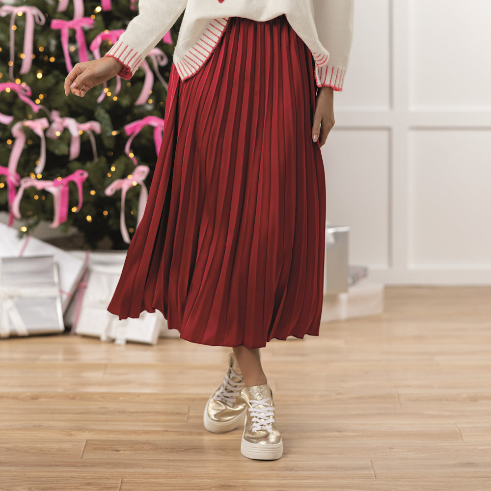 A person wearing a red Townes pleated maxi skirt with a white sweater and gold sneakers by a festive Christmas tree.