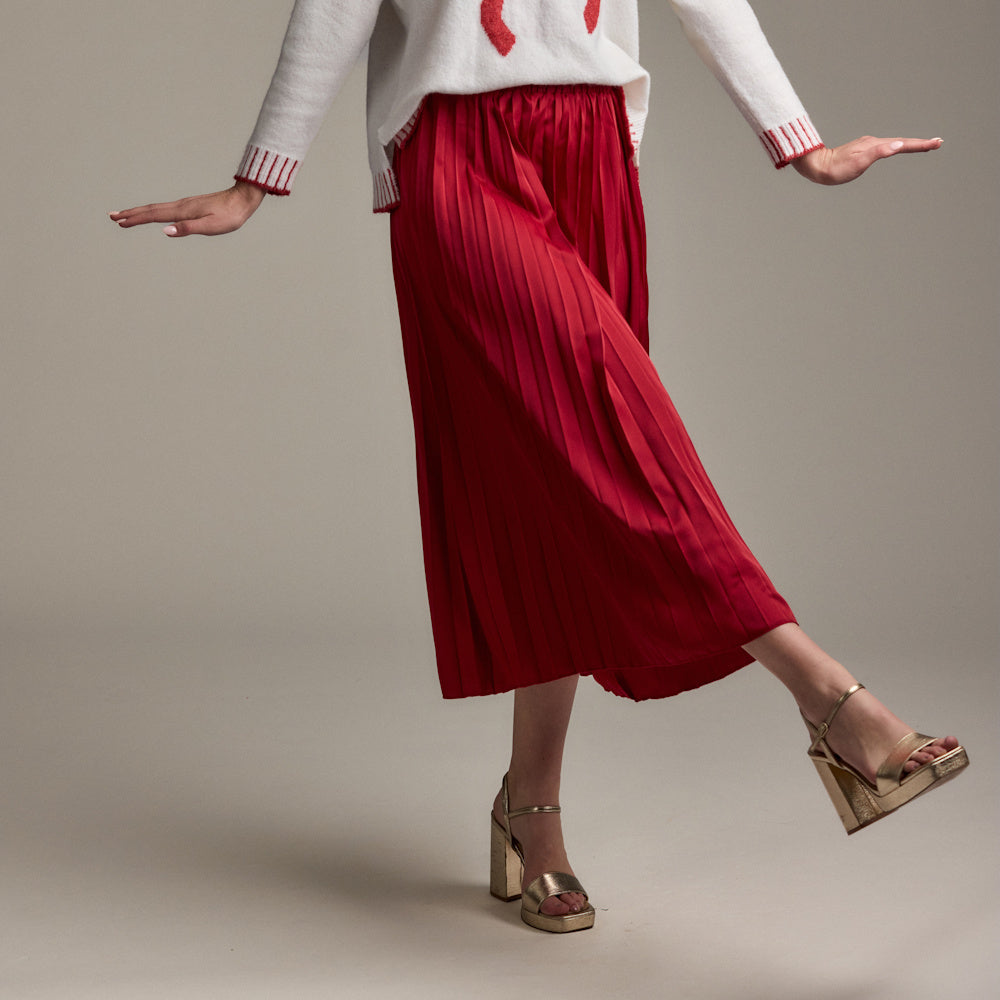 A woman wears the pleated, midi-length Townes skirt in red with gold block heels and a white sweater against a beige background.