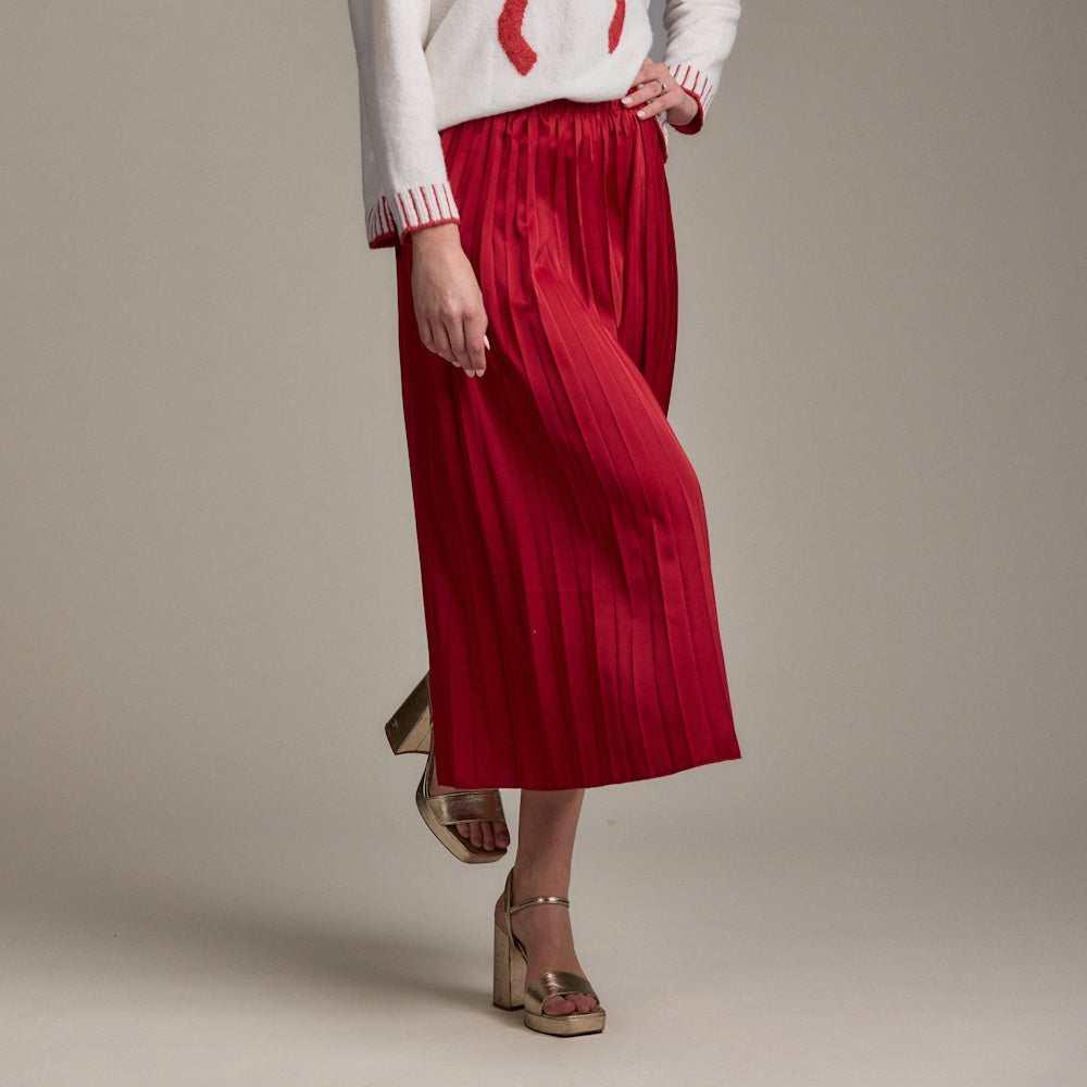 A woman in a red Townes pleated maxi skirt and a white sweater poses, showcasing a pair of metallic gold platform sandals.