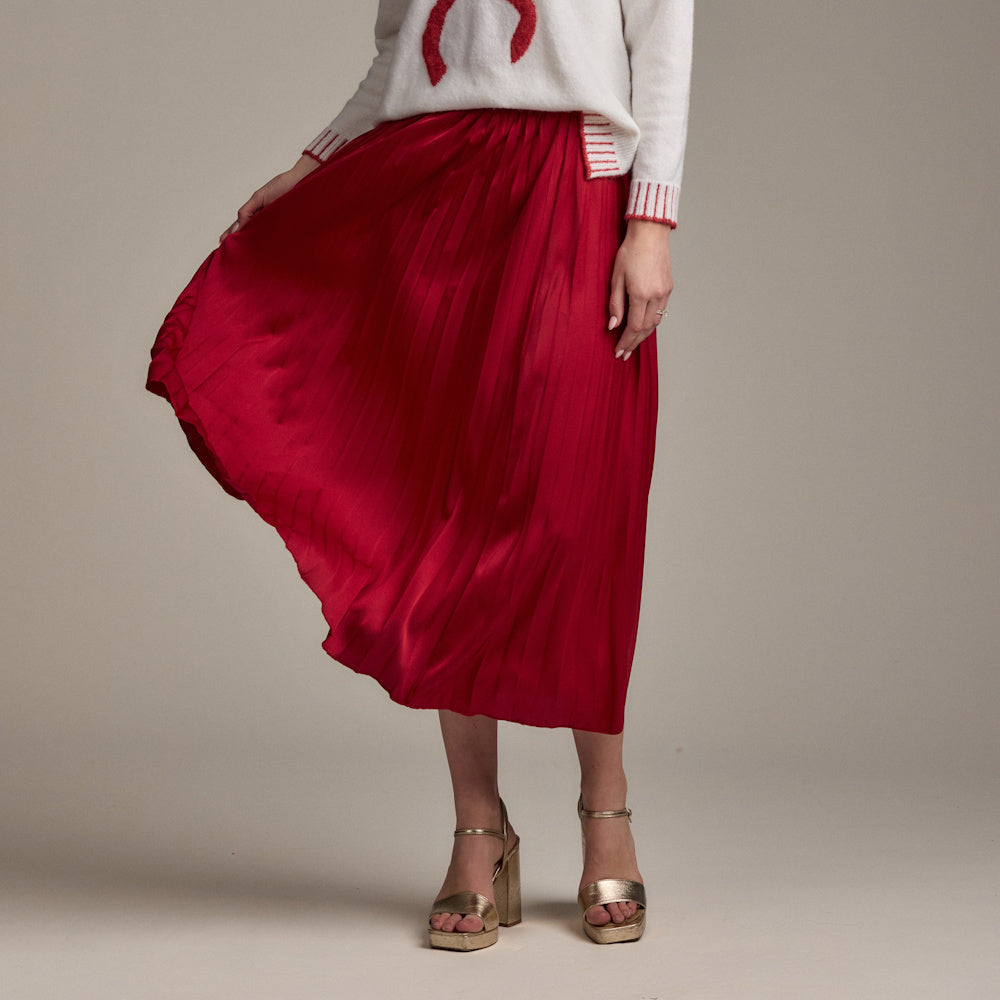 A woman models a red pleated midi skirt with a white graphic sweater and gold platform heels against a gray background.