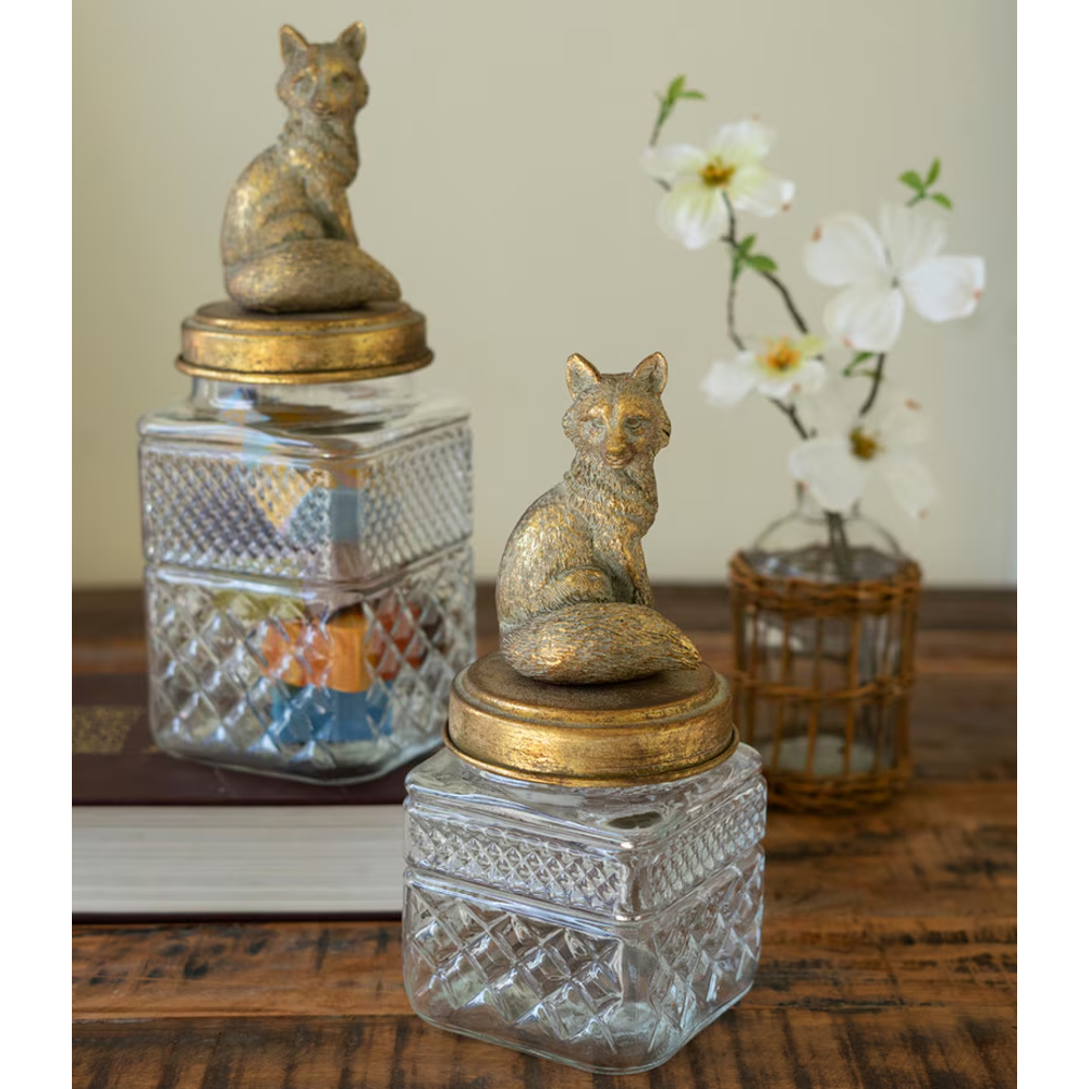 Textured glass canisters with brass fox lids styled on a wooden tabletop with flowers in a vase.
