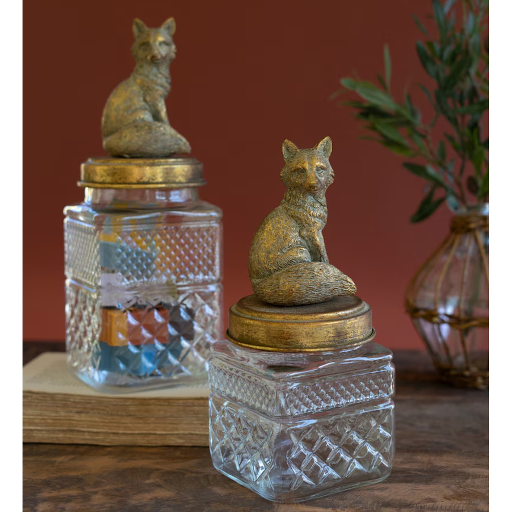 Two diamond-patterned glass canisters with brass fox lids sitting on a book and wooden table.