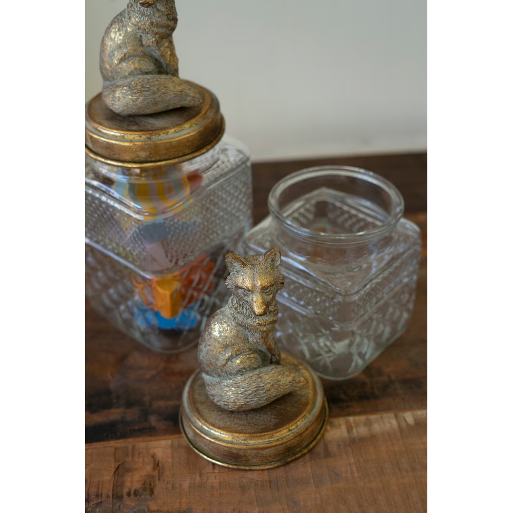 Top-down view of textured glass canisters with brass fox lids, with one lid set on the wooden table.