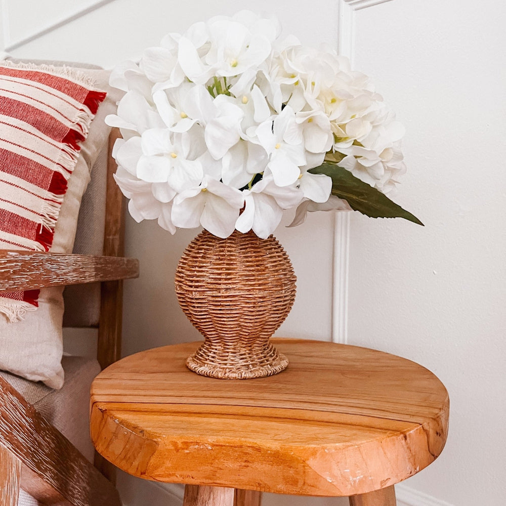 Woven resin basket vase with white hydrangea flowers on a rustic wooden side table next to a red striped pillow.