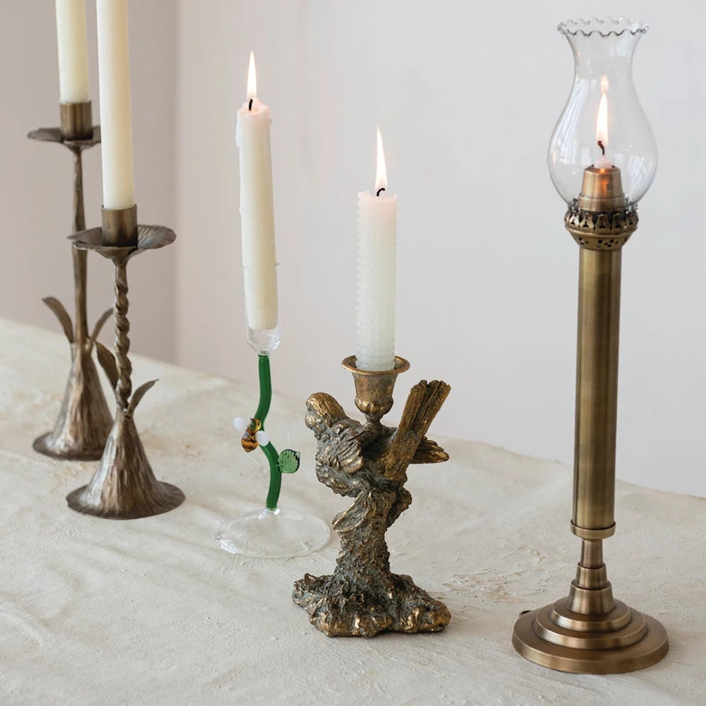 Vintage gold bird taper holder and assorted brass candlesticks with lit white candles on a cream tablecloth.