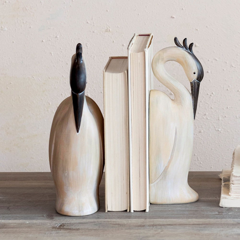 Heron-shaped resin bookends holding up two cream books on a rustic wooden shelf.