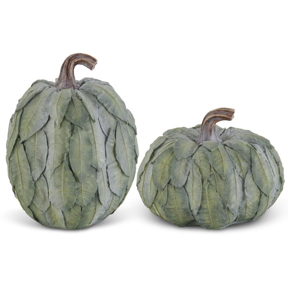Two decorative sage green pumpkins covered in layered leaves with curved brown stems on a white backdrop.