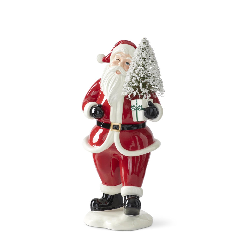 Traditional winking Santa Claus figurine in a red suit holding a gift box and mini snowy bottle brush tree.