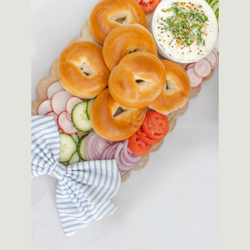 Bagel brunch board with cream cheese, sliced radishes, cucumbers, and red onions on a scalloped wood tray with a blue bow.