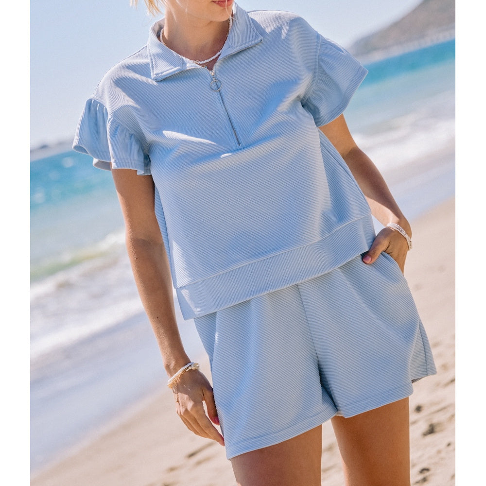 A person wearing a light blue ribbed short-sleeve quarter-zip top and matching shorts on a sandy beach.