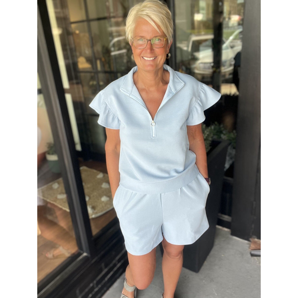 A smiling woman wearing a light blue matching short set with ruffled sleeves and a quarter-zip top.