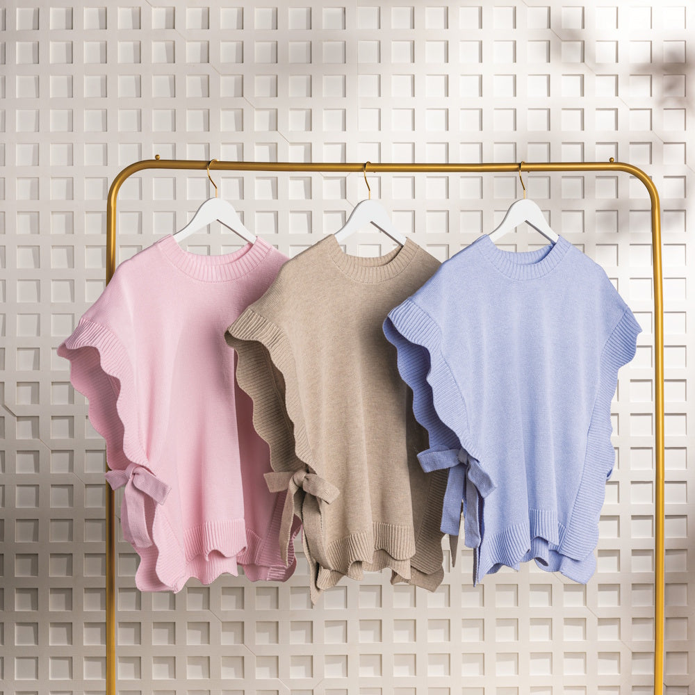 Three short sleeve knit sweater in pink, beige, and blue with ruffled sides and bow details hang on a gold clothing rack.