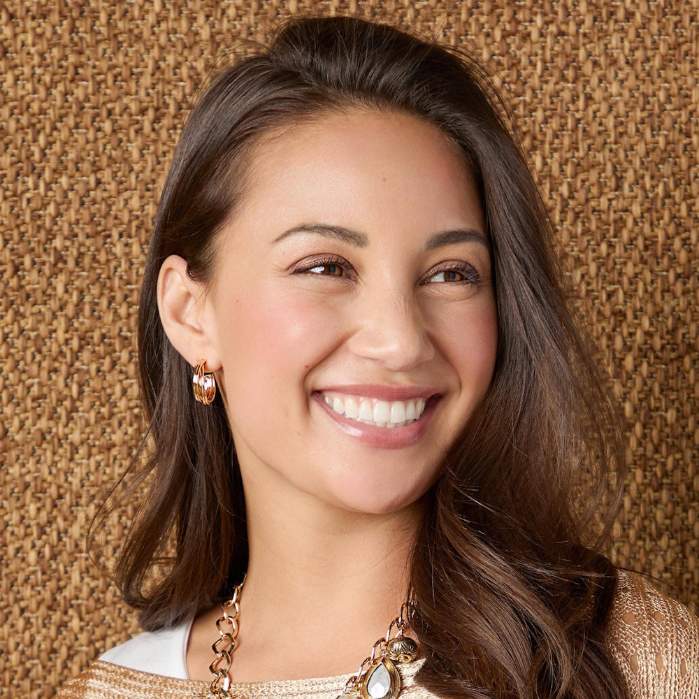 Woman smiling wearing gold stacked hoop earrings and a gold chain necklace against a textured brown background.