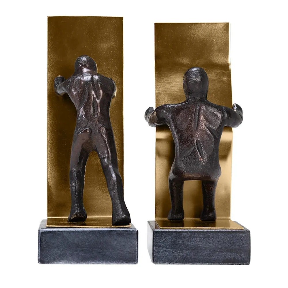 Two sculpted figures leaning against gold metal bookends, viewed from behind on dark stone bases.