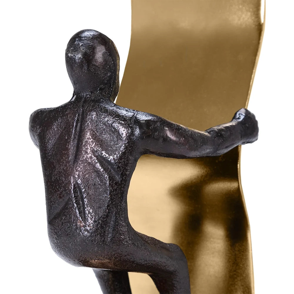 A dark textured metallic sculpture of a man leaning against a curved gold metal surface, viewed from behind.