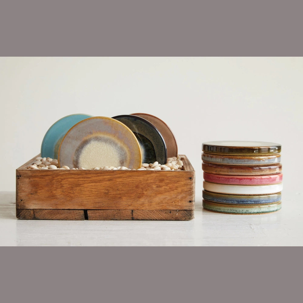 Colorful glazed round stoneware trivets stacked and displayed in a wooden box with white pebbles.