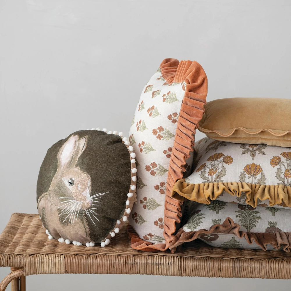 Round rabbit pillow resting on a woven bench beside a stack of patterned decorative throw pillows.