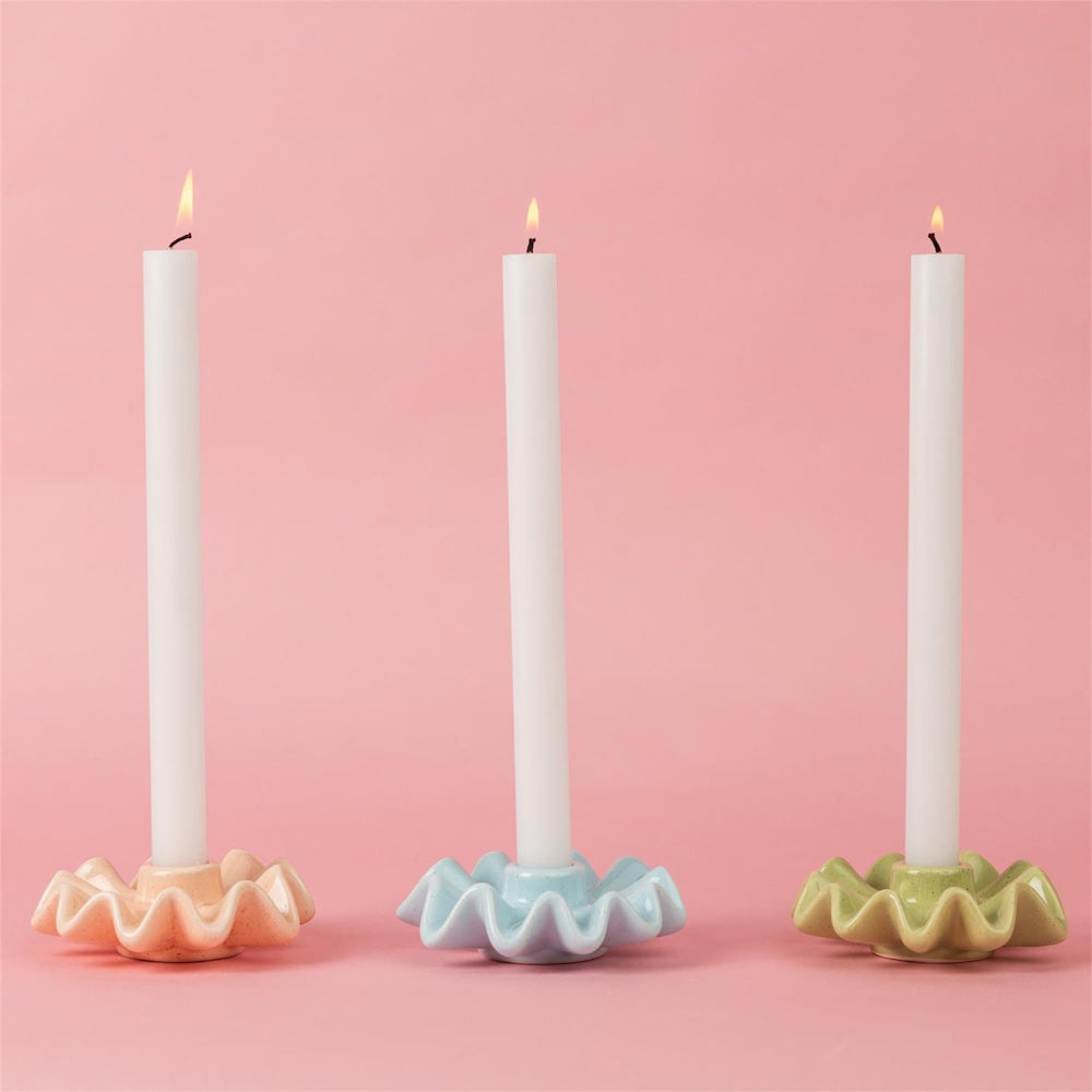 Three lit white taper candles in pastel ruffled ceramic holders against a pink background.