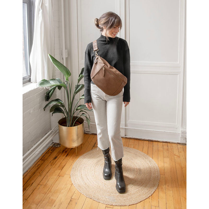 Woman wearing a russet brown large sling crossbody bag over a black sweater and white jeans in a bright room.