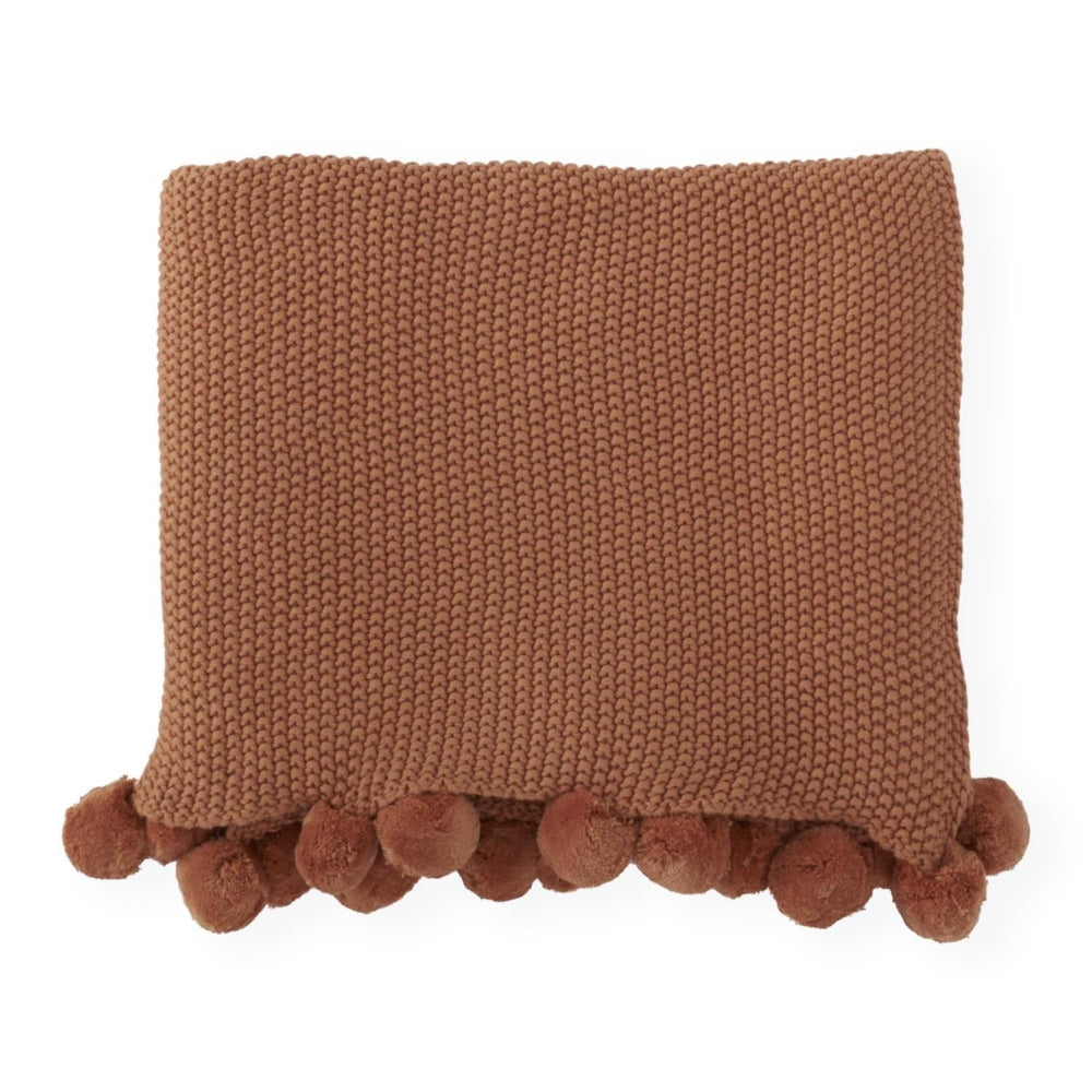 A neatly folded rust-brown moss-stitch knit throw blanket featuring a playful pom-pom trim on a white background.