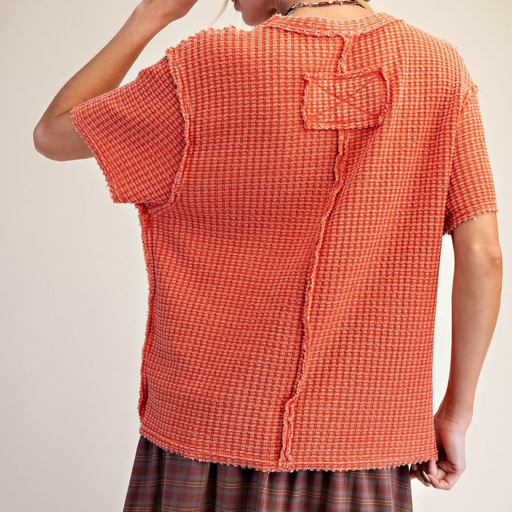 Back view of a person wearing a rust-orange waffle-knit top with raw edge seams and a patch detail.