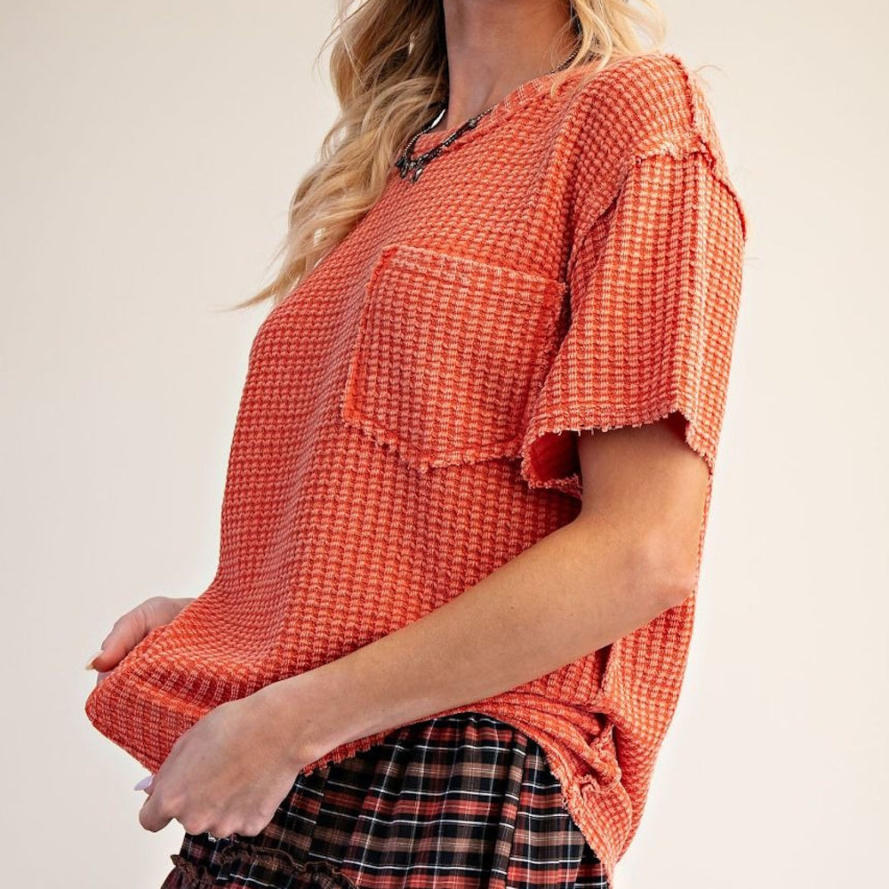 Rust-colored waffle-knit short-sleeve top with raw edges and a front chest pocket, shown from a side angle.