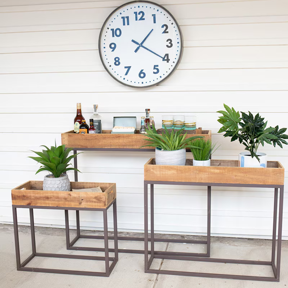 Three rustic wooden tray console tables on dark metal bases styled with plants and bar supplies.