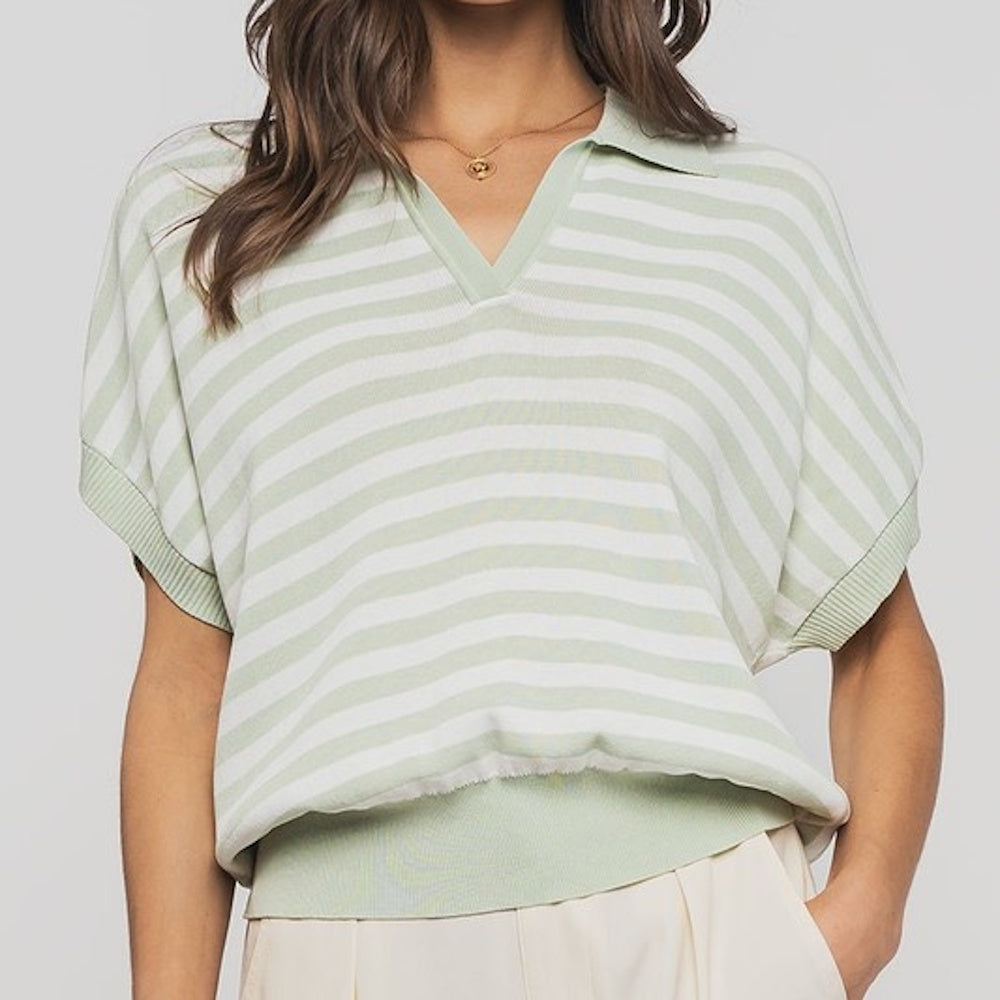 Woman wearing a striped sage green and white V-neck polo knit top with ribbed trim and light-colored trousers.