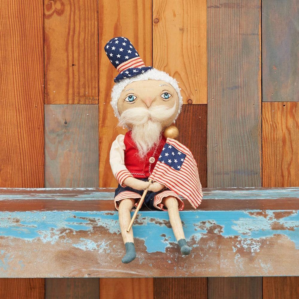 A seated fabric doll with white hair, a tall stars-and-stripes hat, and a red vest holding a small American flag.