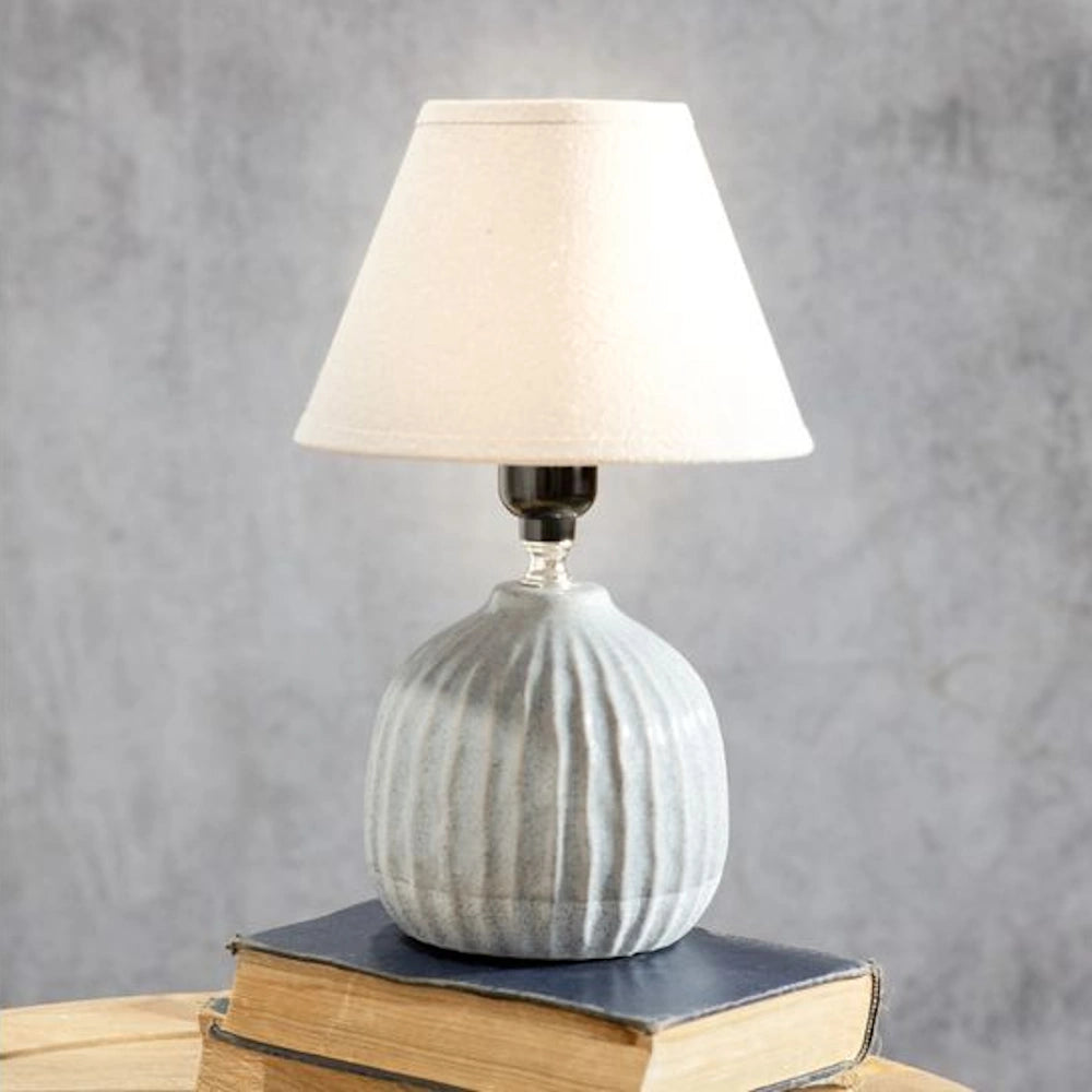 Small ceramic table lamp with a ribbed grey base and white fabric shade sitting on a stack of books.