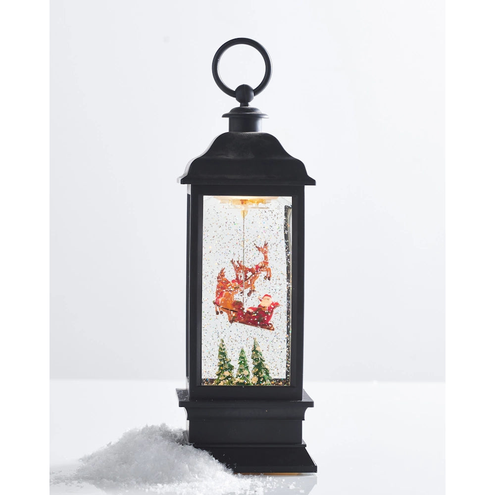 A lighted black water lantern with Santa in his sleigh flying over trees, accented by a pile of artificial snow at the base.