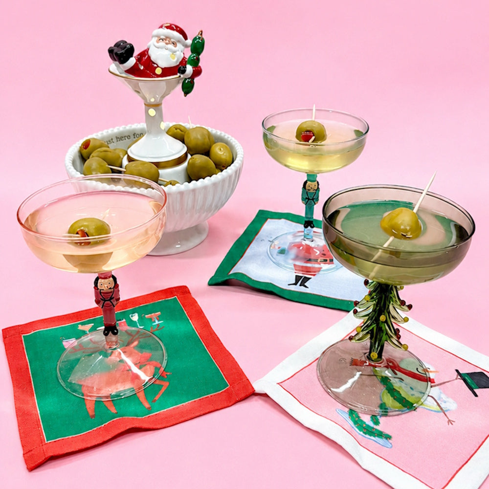 Holiday cocktail glasses with festive stems served alongside a Santa-themed olive bowl on a pink surface.