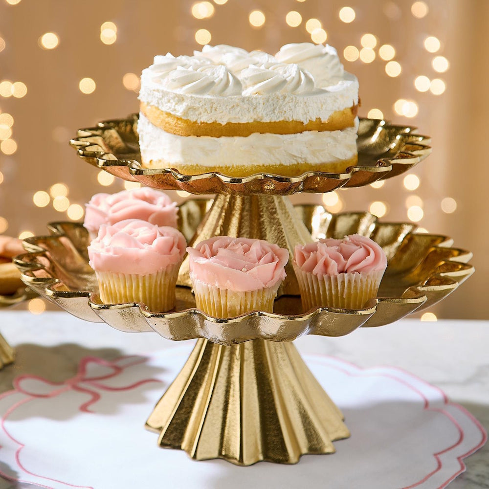 Gold tiered cake stand holding a white cake and pink cupcakes in front of warm bokeh lights.
