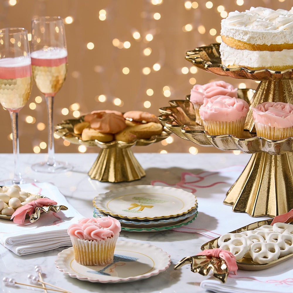 Gold cake stands, pink cupcakes, cake, and champagne on a festive dessert table with warm bokeh lights.