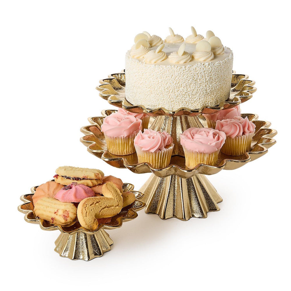 Gold cake stands displaying a white layered cake, pink frosted cupcakes, and assorted cookies.