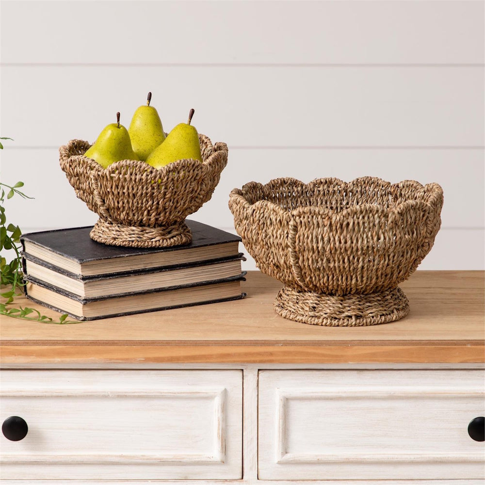 Two woven seagrass baskets on a wooden tabletop; one sits on a stack of books and holds three green pears.