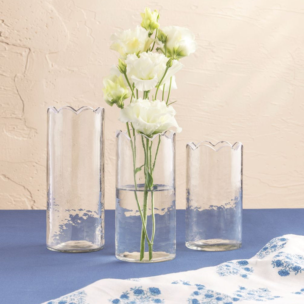 Three scalloped glass bud vases in varying heights with white lisianthus flowers on a blue tabletop and textured wall.