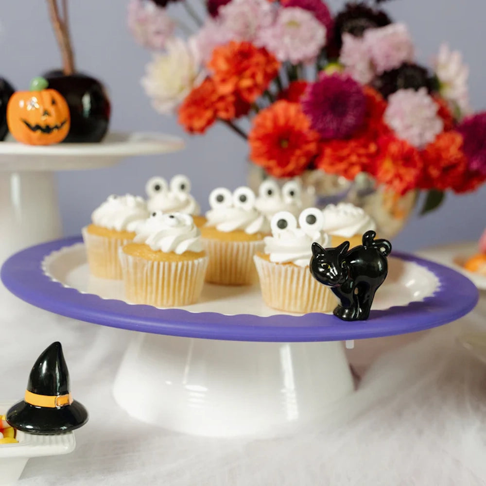 A purple-rimmed cake stand holds monster cupcakes with candy eyes and a small, arched black cat figurine.