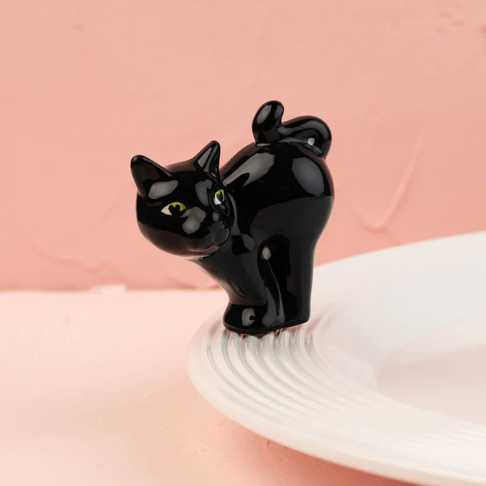 A glossy black cat figurine with arched back and yellow eyes, perched on the rim of a white plate.