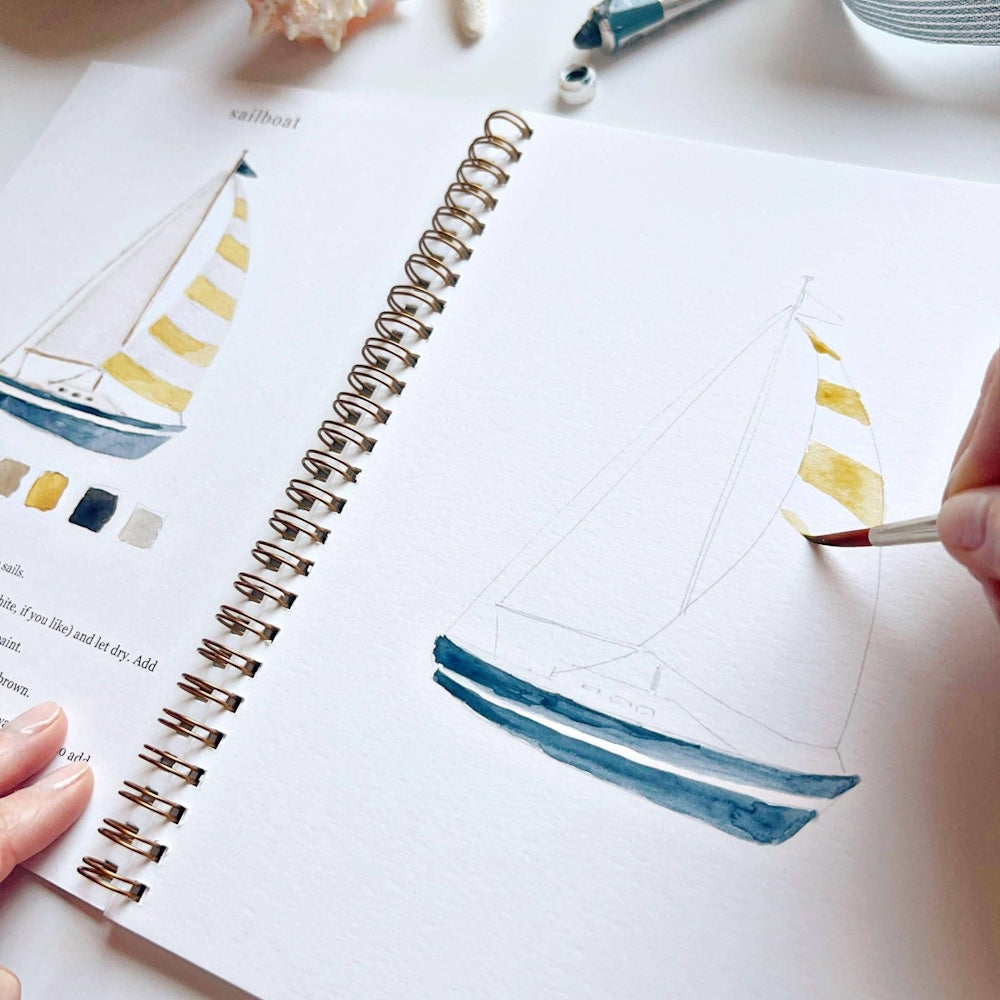 Watercolor sailboat tutorial in a spiral workbook with yellow and blue paint being applied by a hand.