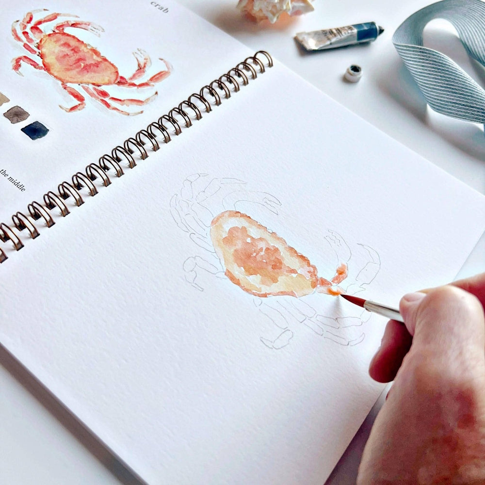 Watercolor painting of a crab in a spiral sketchbook with a paintbrush, paint tube, and blue ribbon nearby.