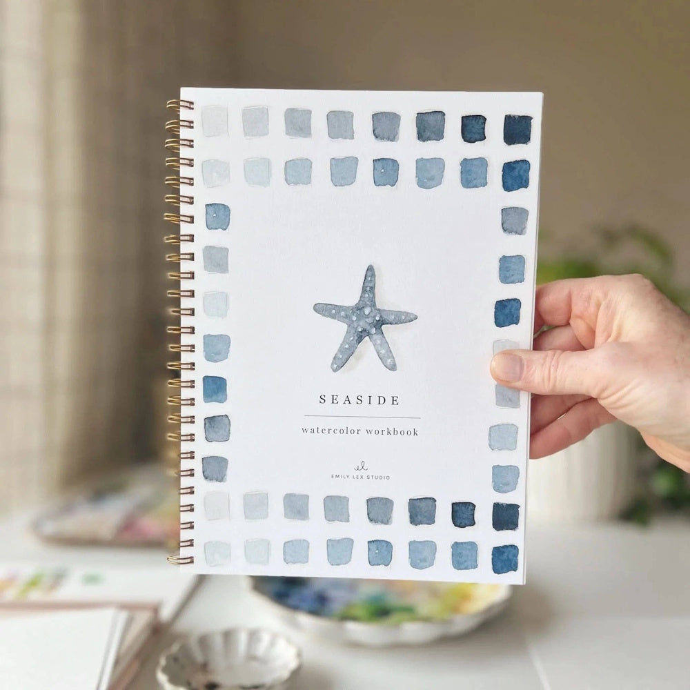 Hand holding Seaside Watercolor Workbook with blue watercolor swatches and starfish illustration by Emily Lex.