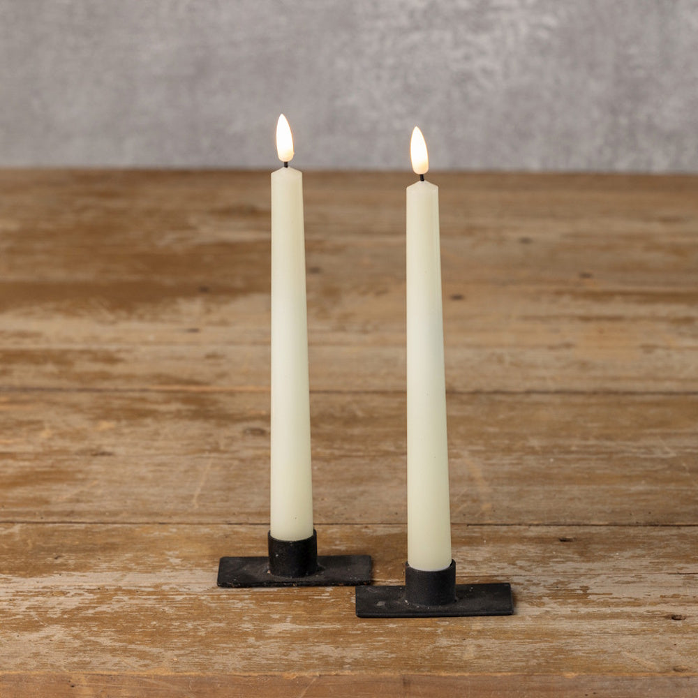 Two lit cream-colored flameless taper candles in black metal holders, standing on a wooden surface.