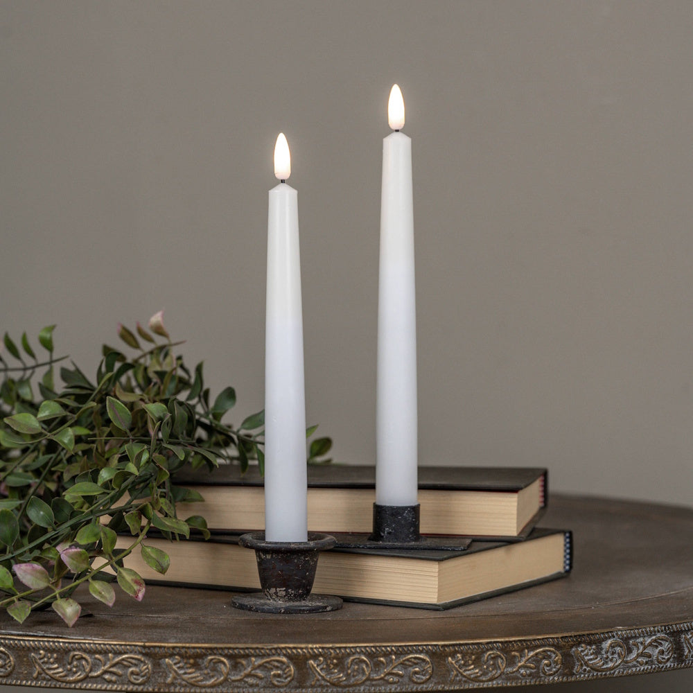 Two white flameless taper candles in dark metal holders on stack of books with greenery on table.
