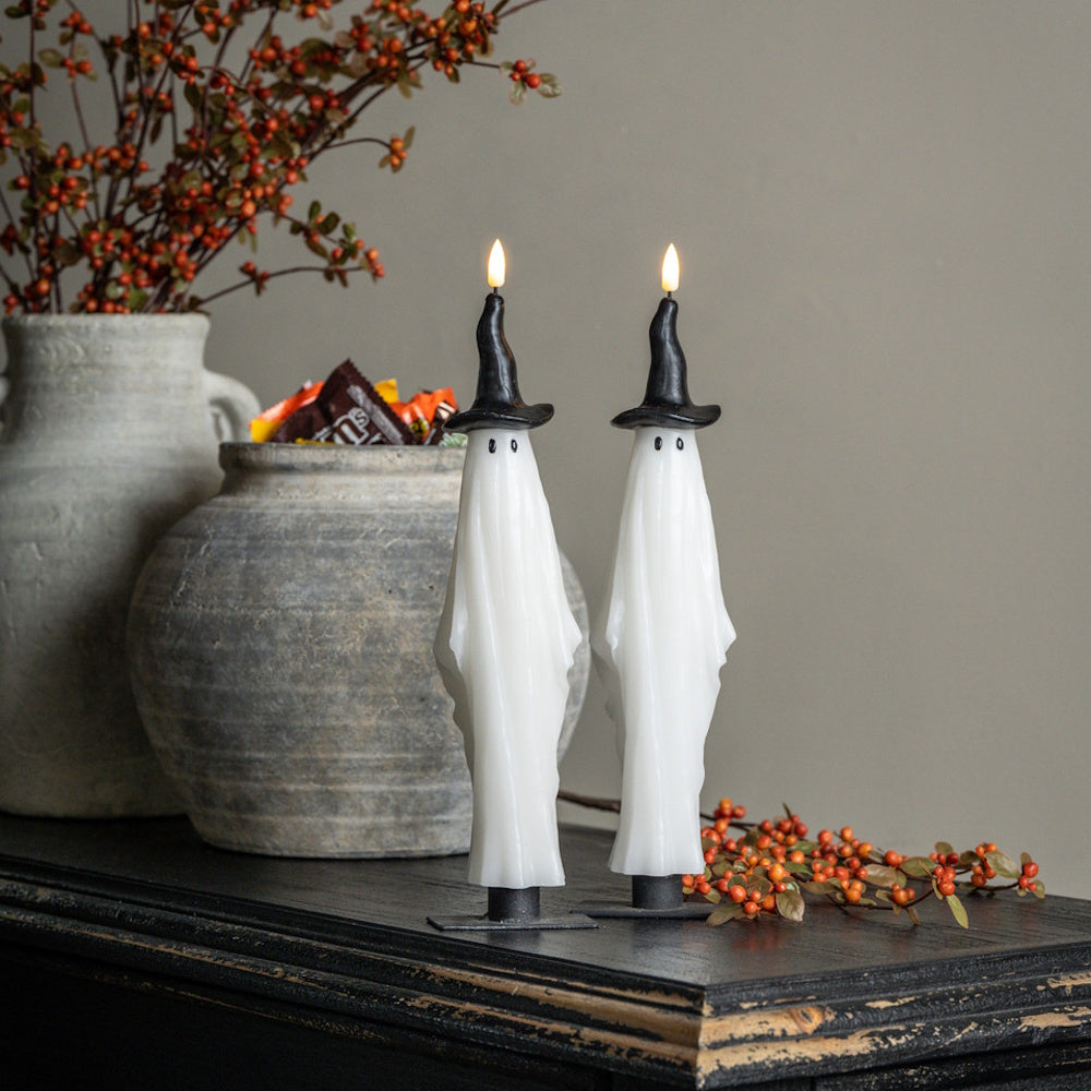 Two white ghost taper candles wearing black witch hats glow atop a dark wooden shelf with festive decor.