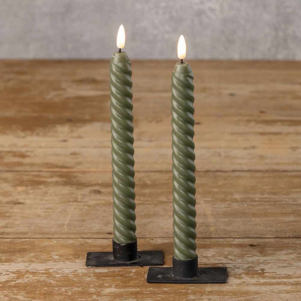 Two lit dark green twisted taper LED candles in simple black metal holders standing on a wooden surface.