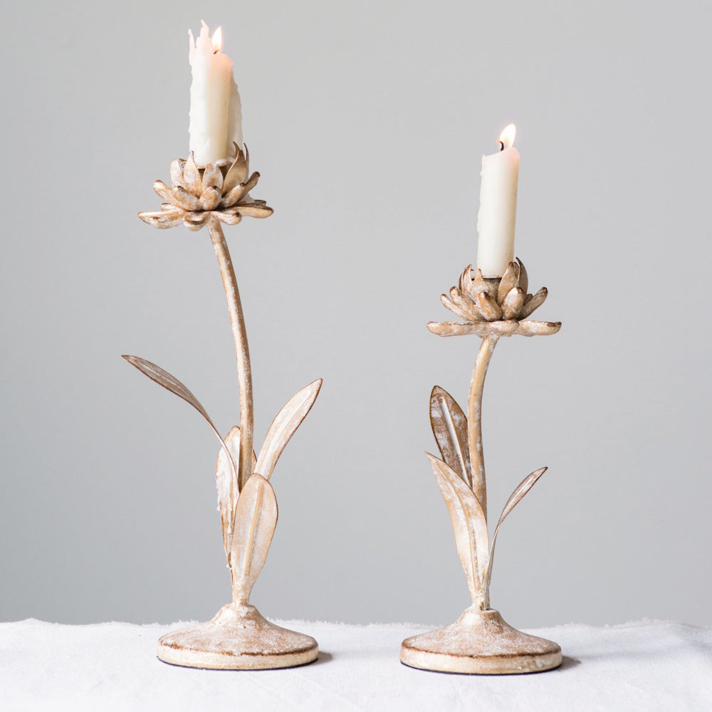 Two metal candle holders shaped like flowering stems with lit white taper candles, on a white surface.
