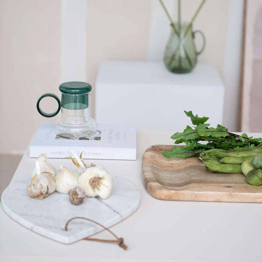 A glass pitcher with a green lid sits on a book next to garlic, a wooden tray, and fresh greens.
