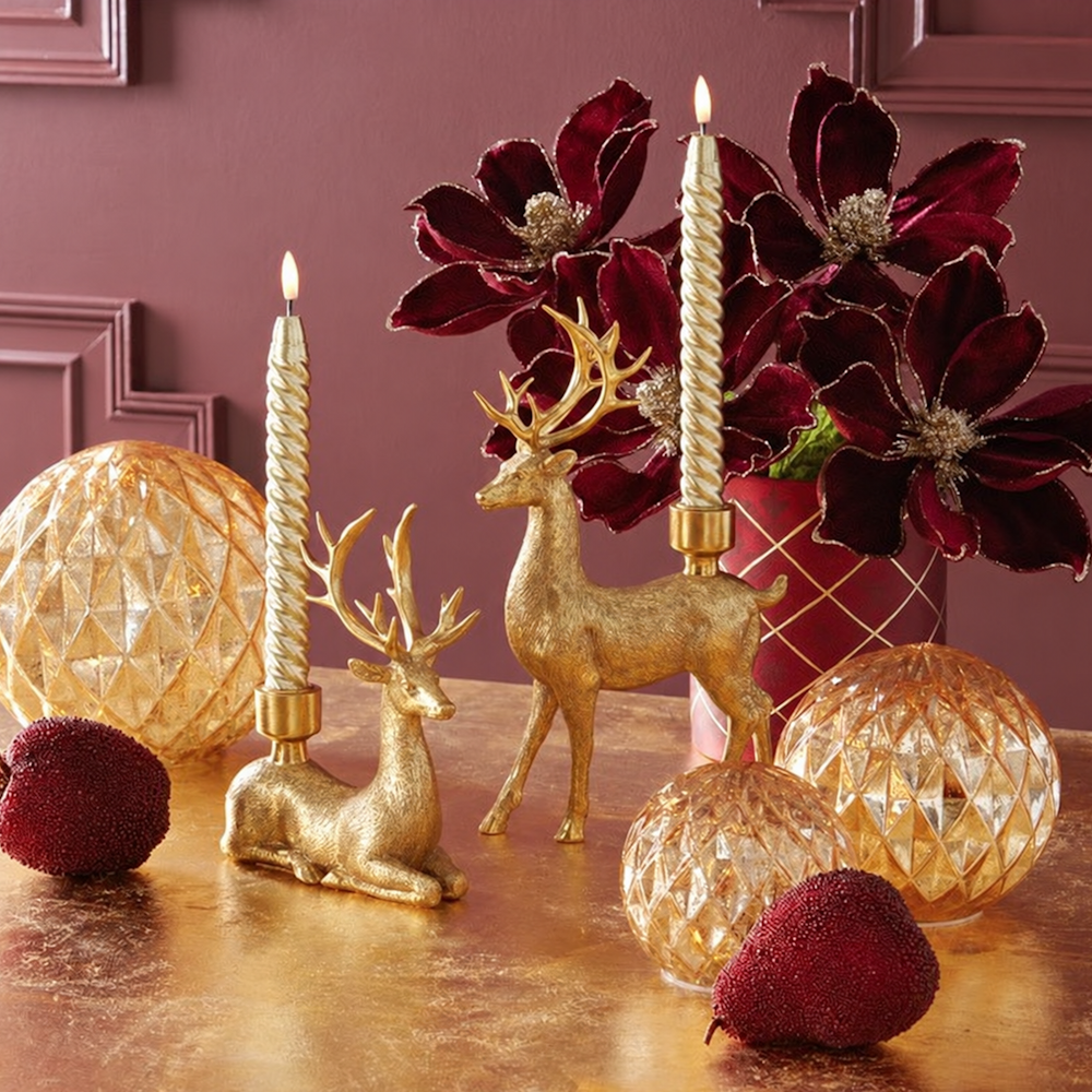 Holiday display featuring gold deer taper candleholders with lit candles, dark red flowers, and gold globes.