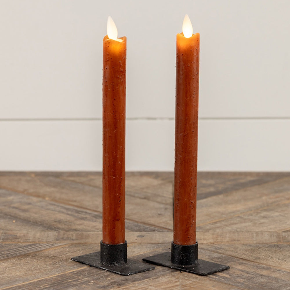Two glowing speckled bronze taper candles in black metal holders standing on a rustic wooden table.